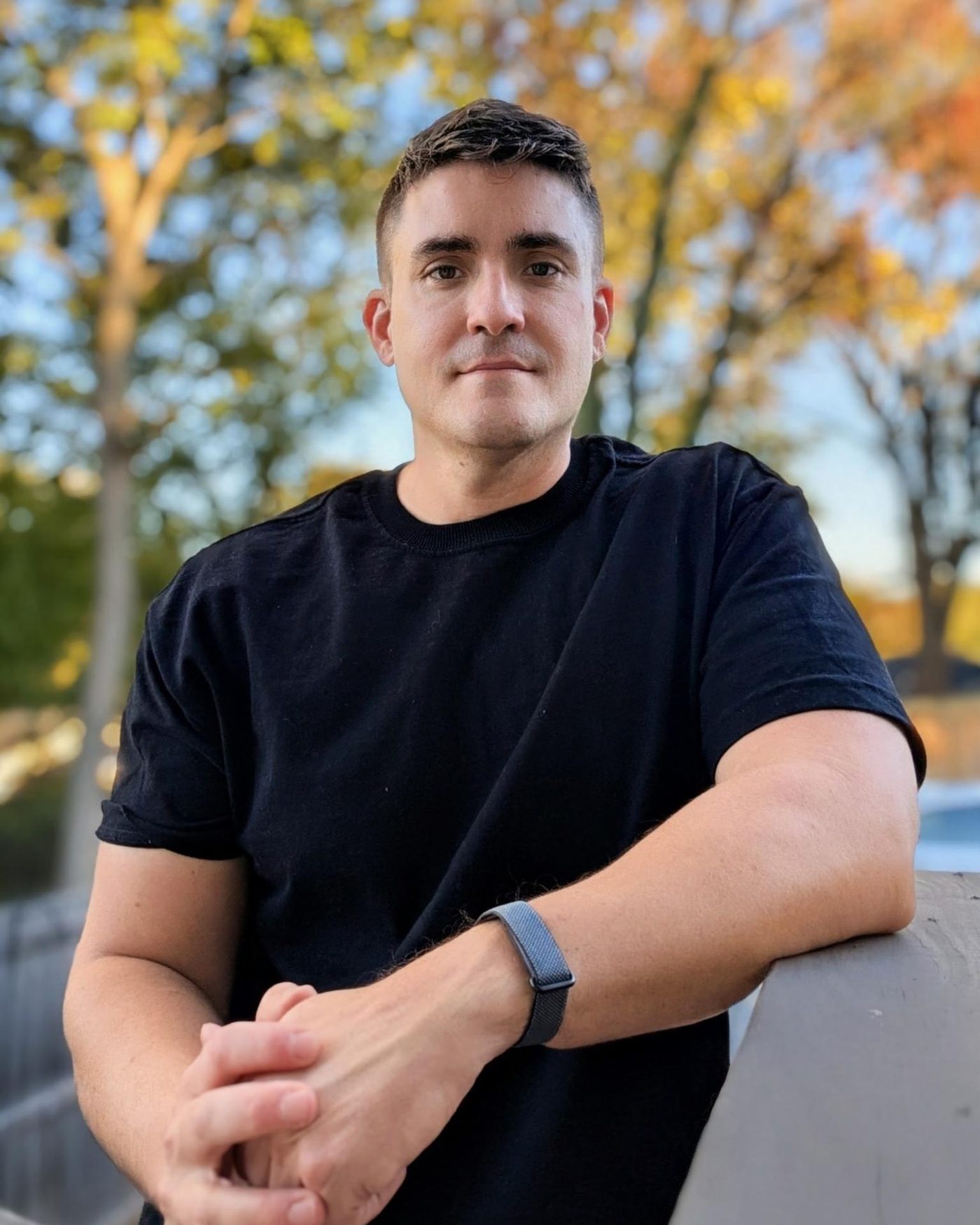Adam Nicholson leaning on a railing outdoors, autumn trees behind him